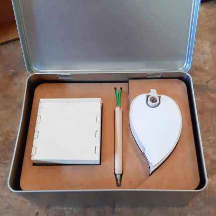 Caja metálica con la tapa abierta y una pieza de cartón dentro en donde están encajados una libreta y un bolígrafo